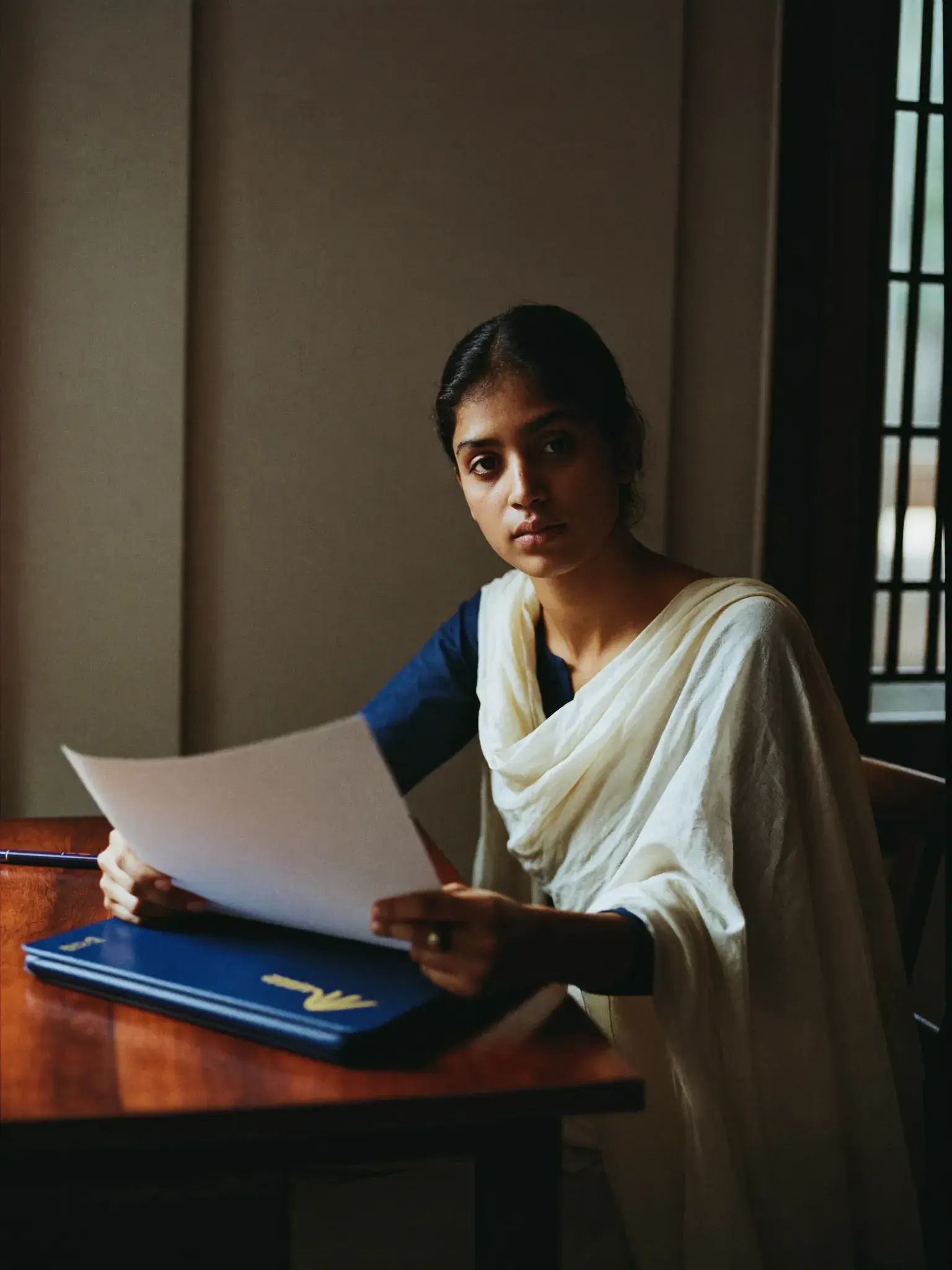 A young woman at a desk by a window, reading through a printed application form.